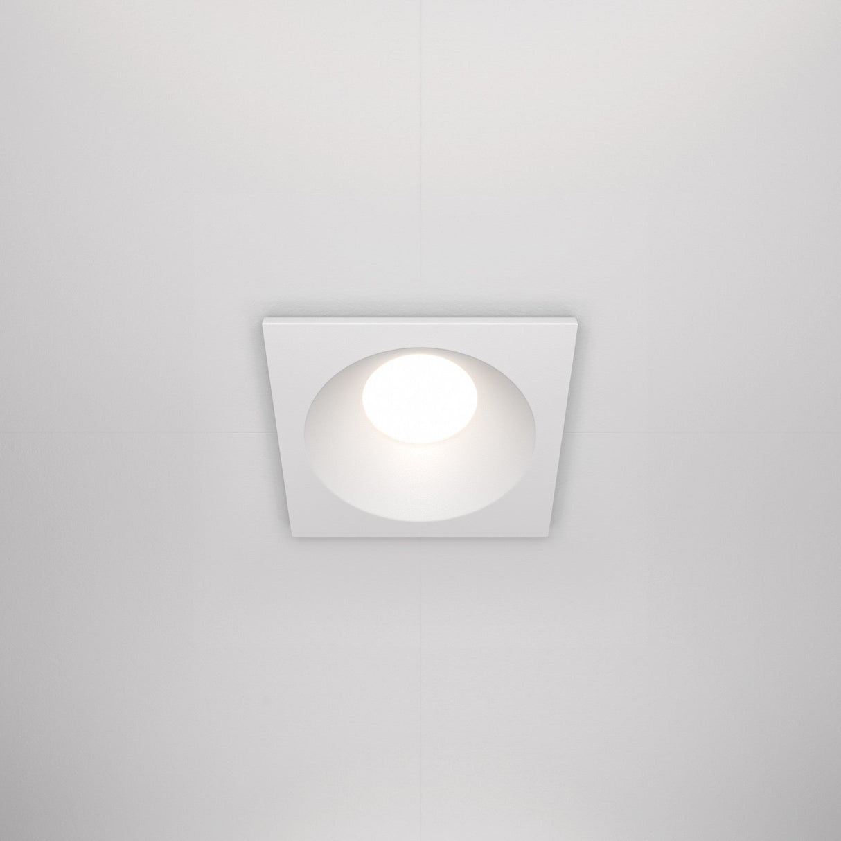 The Zoom Single Fixed Square Recessed Downlight - White offers IP65 rating and emits soft light against a white ceiling. Its minimalist design features a bright circular bulb, blending seamlessly with modern ceiling aesthetics.