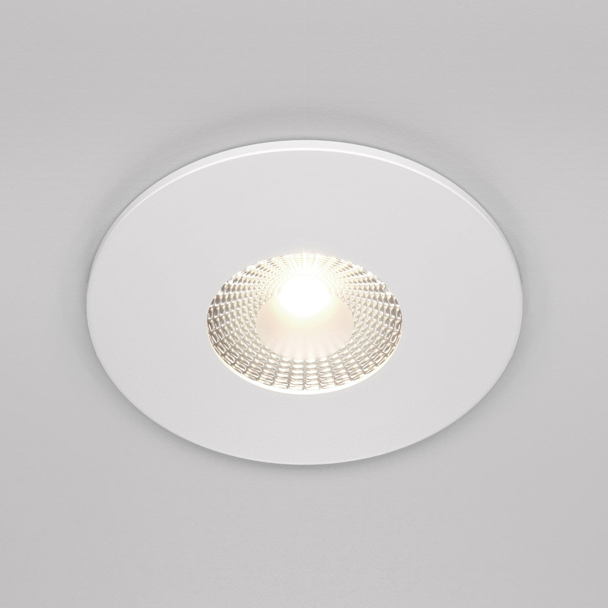 The Zen LED Round Recessed Downlight 7W 4000K - White fixture, turned on, illuminates its textured interior and casts a warm glow on the neutral ceiling, showcasing energy-efficient lighting.