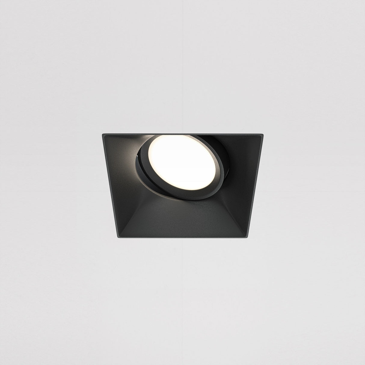 The Dot Single Tilt Square Plaster-in Recessed Downlight in black features a modern design with a circular LED bulb emitting bright white light against a plain white ceiling, offering an elegant and energy-efficient lighting solution.