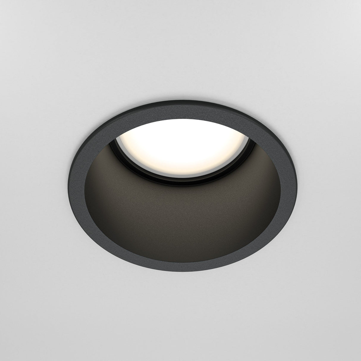 Close-up of the Reif Round Recessed Downlight - Black, featuring a matte black finish. It emits a soft white glow and is elegantly centered on a plain white ceiling as ambient lighting.