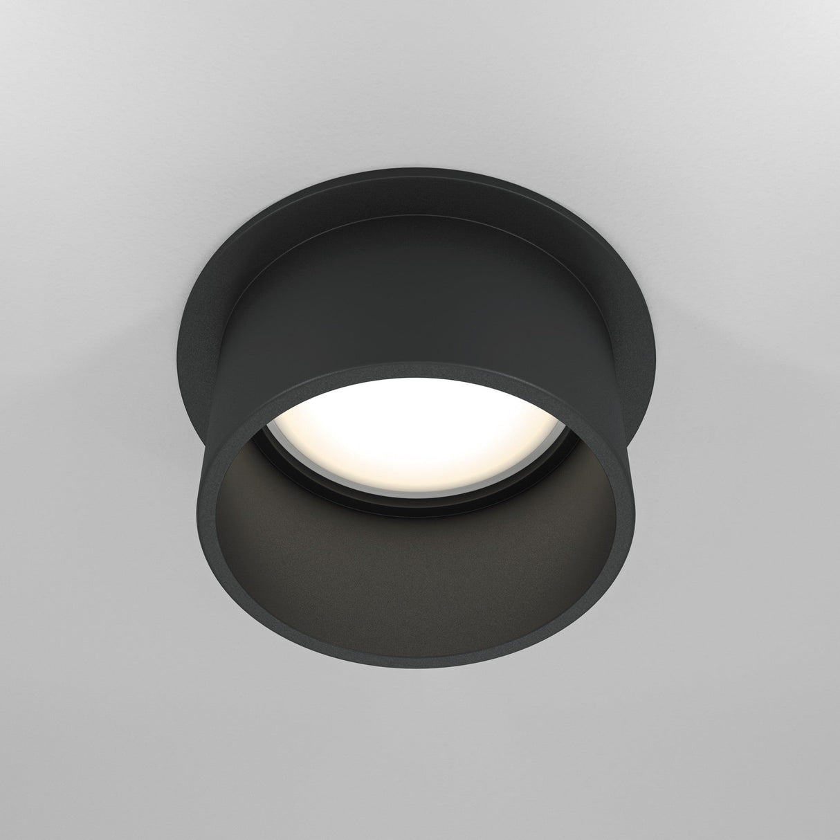 The Reif Round Recessed Downlight in black offers a minimalist, cylindrical design with an extended rim. It casts a soft, warm glow and blends seamlessly with the white ceiling.
