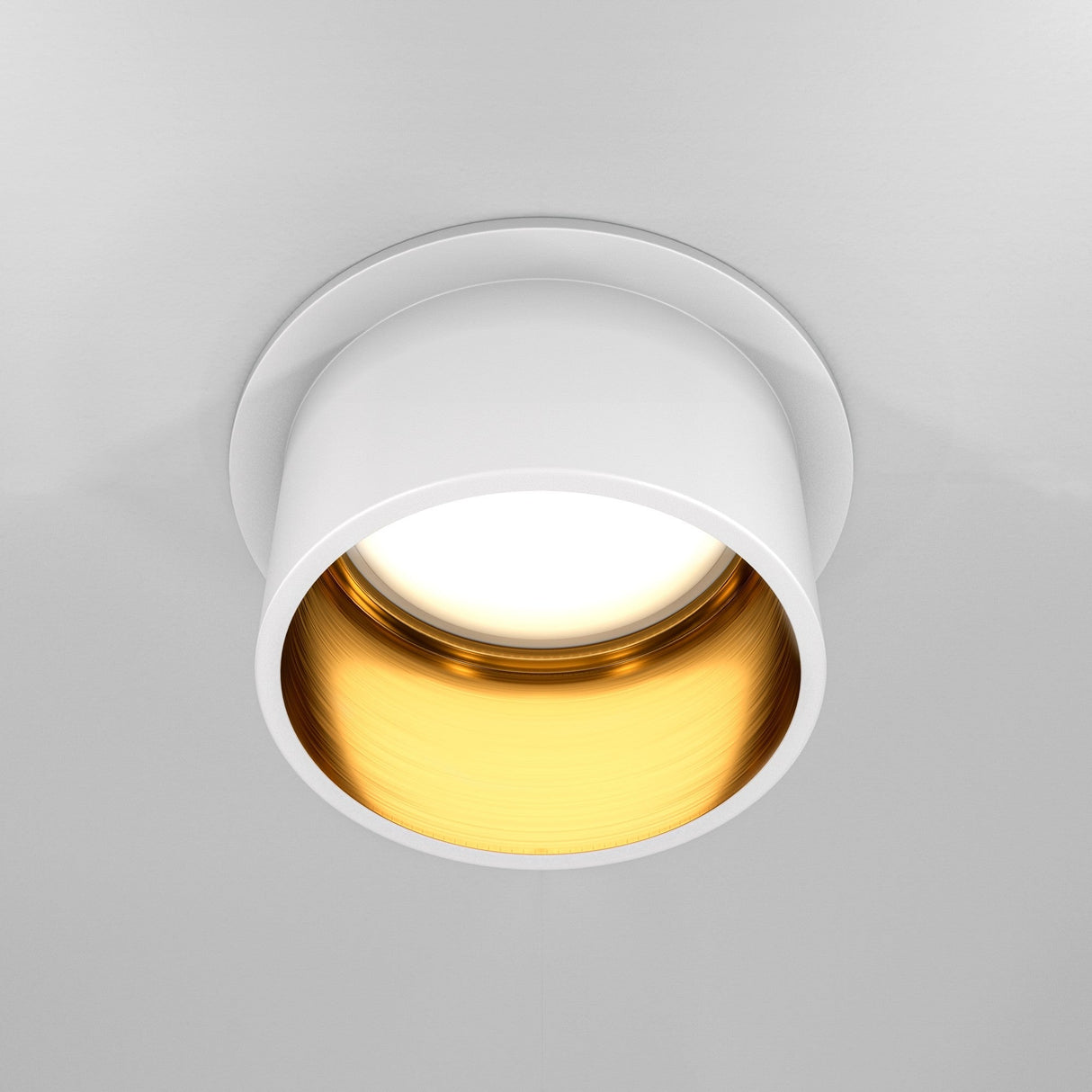 The Reif Round Recessed Downlight, featuring a white & gold finish with an extended rim, is mounted on a plain white ceiling and emits a soft glow from its gold-toned interior.