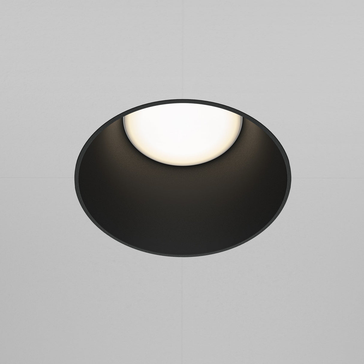 The Share Round Plaster-in Fixed Recessed Downlight in black emits a soft white glow, enhancing minimalist lighting. It is elegantly installed within a white ceiling with subtle grid lines.
