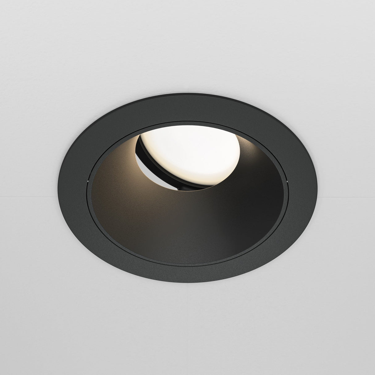 The Share Round Recessed Swivel Downlight in black is embedded in a white ceiling and features an off-white bulb. It has a swivel mechanism that allows the bulb to angle inward, creating a soft accent glow against the fixture.