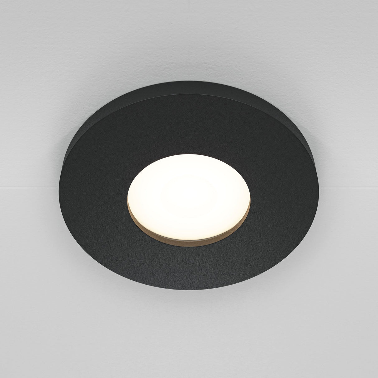 The Stark Fixed Round Recessed Downlight in black elegantly mounts on a white ceiling, featuring a dark outer ring and bright, glowing center to cast soft illumination. This stylish fixture enhances any space with its modern design.