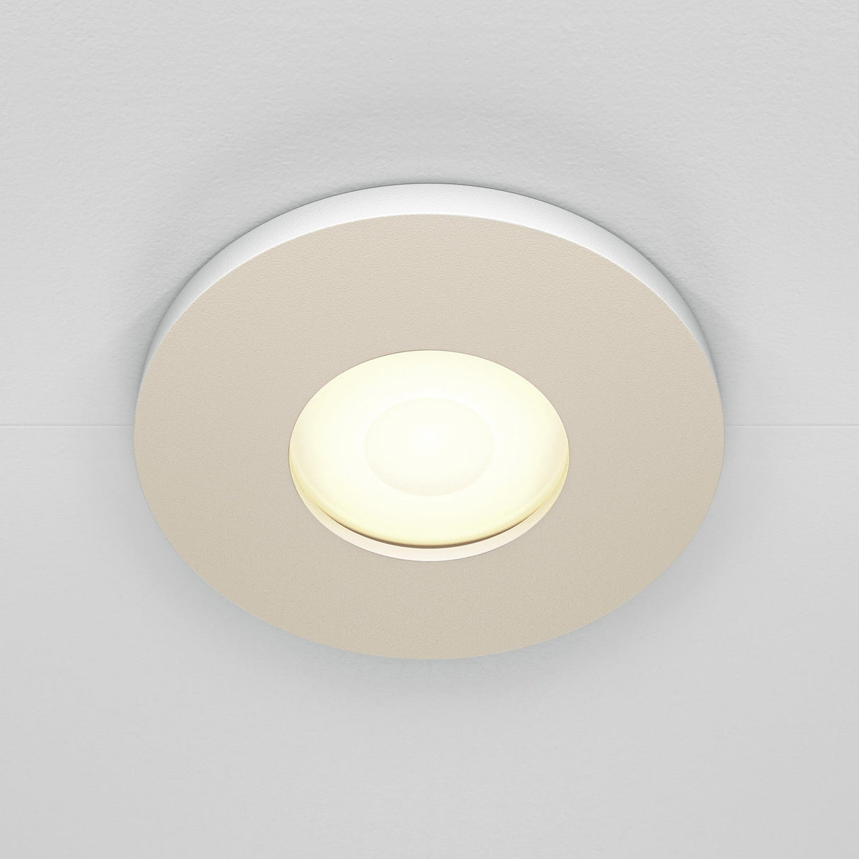 The Stark Fixed Round Recessed Downlight - White, featuring a glowing GU10 bulb, is set in a smooth surface. Its modern design emits soft, warm light and boasts an IP65 rating for dependable use in moist areas.