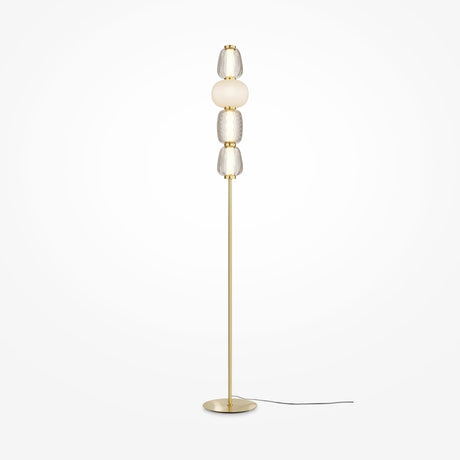 The Pattern LED Floor Lamp - Gold is tall and modern with a slim gold base and pole, featuring four stacked textured glass orbs in clear and frosted finishes. Its black power cord enhances energy efficiency against a plain white backdrop.