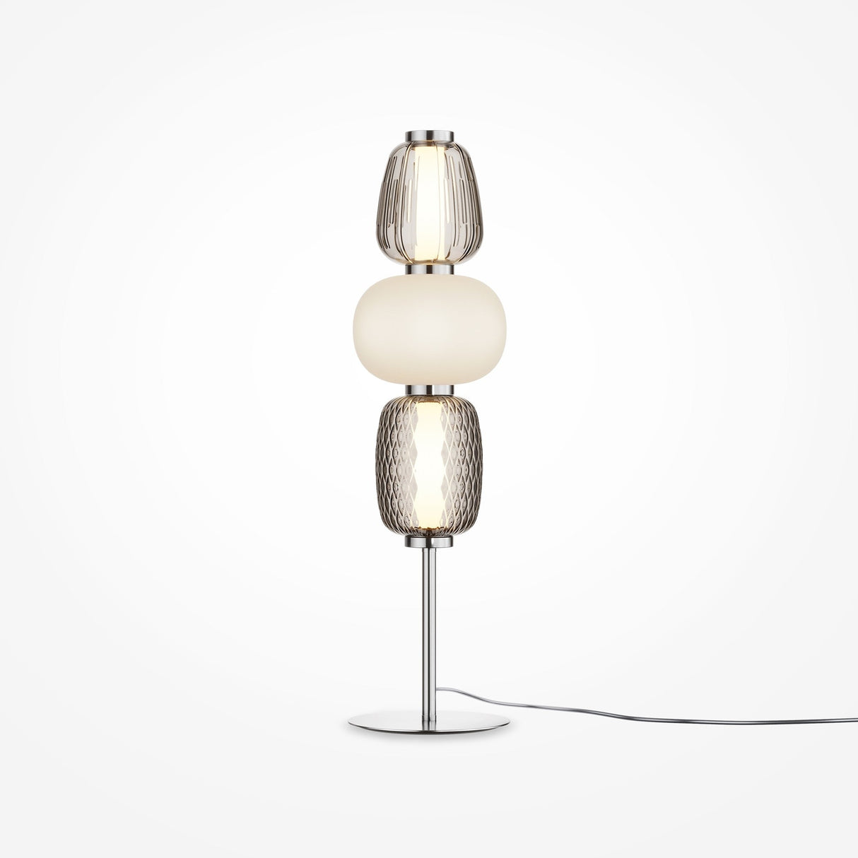 The Pattern LED Table Lamp - Chrome offers a modern look with its chrome finish and metallic base, featuring three stacked glass shades: textured clear on top, smooth white in the middle, and textured amber at the bottom. A black power cord enhances its contemporary appeal.