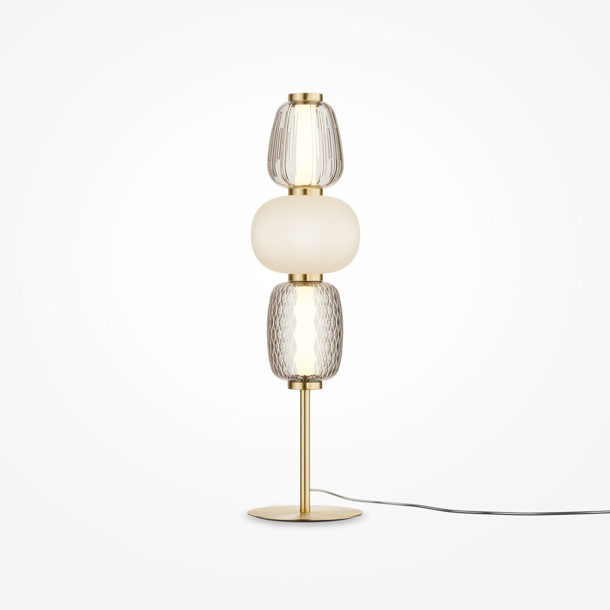 The Pattern LED Table Lamp - Gold features a sleek brass base with gold metal fittings, three stacked glass elements (two clear with textured patterns and one opaque white in the center), and an energy-efficient black cord extending from the base.