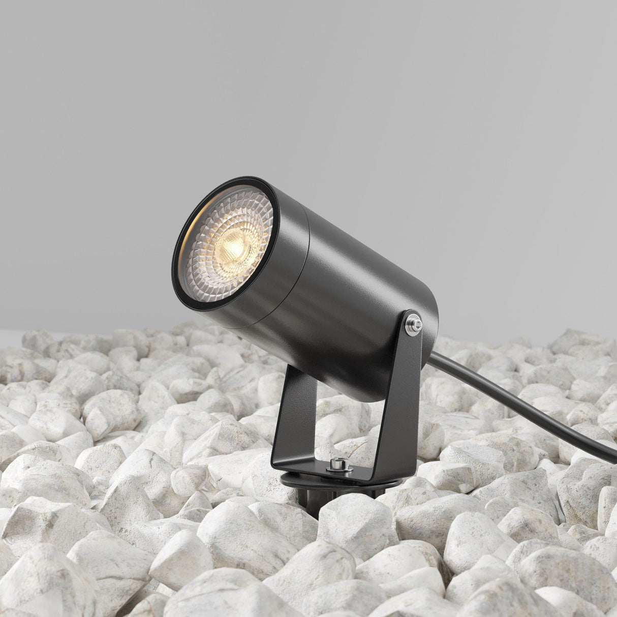 The Scope Outdoor Spike Light - Black, featuring a cylindrical design and adjustable spotlight, is mounted on a base to illuminate white stones with its cable extending behind it against a plain gray backdrop.