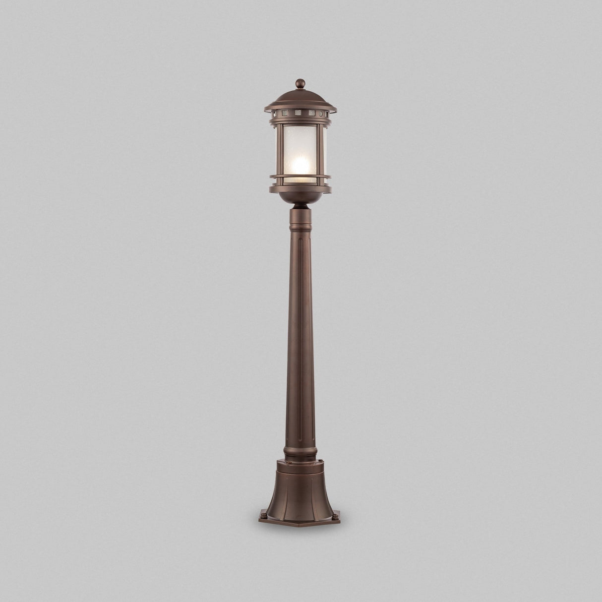 The Salamanca 1 Light Pillar Lantern - Brown features a vintage style with a tall bronze post and cylindrical lantern, emitting soft light through clear glass. Its sturdy and ideal for gardens or pathways against any gray backdrop.