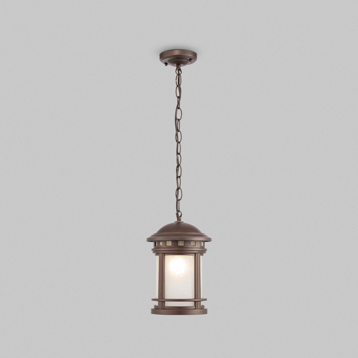 The Salamanca Outdoor Pendant Light - Brown features a lantern-style design hanging from the ceiling by a sturdy chain. It includes a cylindrical frosted glass shade encased in decorative bronze, combining retro aesthetics with durable construction.