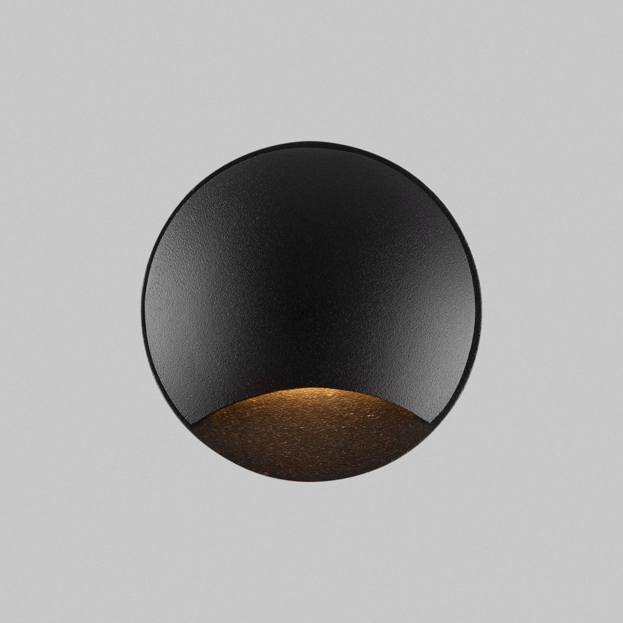 The Biscotti Outdoor 1 Beam LED Recessed Wall Light in black features a minimalist round design, casting a warm downward glow. Its mounted on a light gray surface, emphasizing its modern and sleek look, making it perfect for outdoor lighting in any setting.
