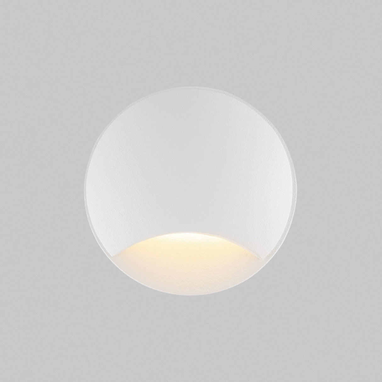 The Biscotti Outdoor 1 Beam LED Recessed Wall Light features a minimalist circular design with a cut-out bottom half, offering energy-efficient and warm illumination. This modern white fixture creates an elegant effect against a plain gray backdrop.