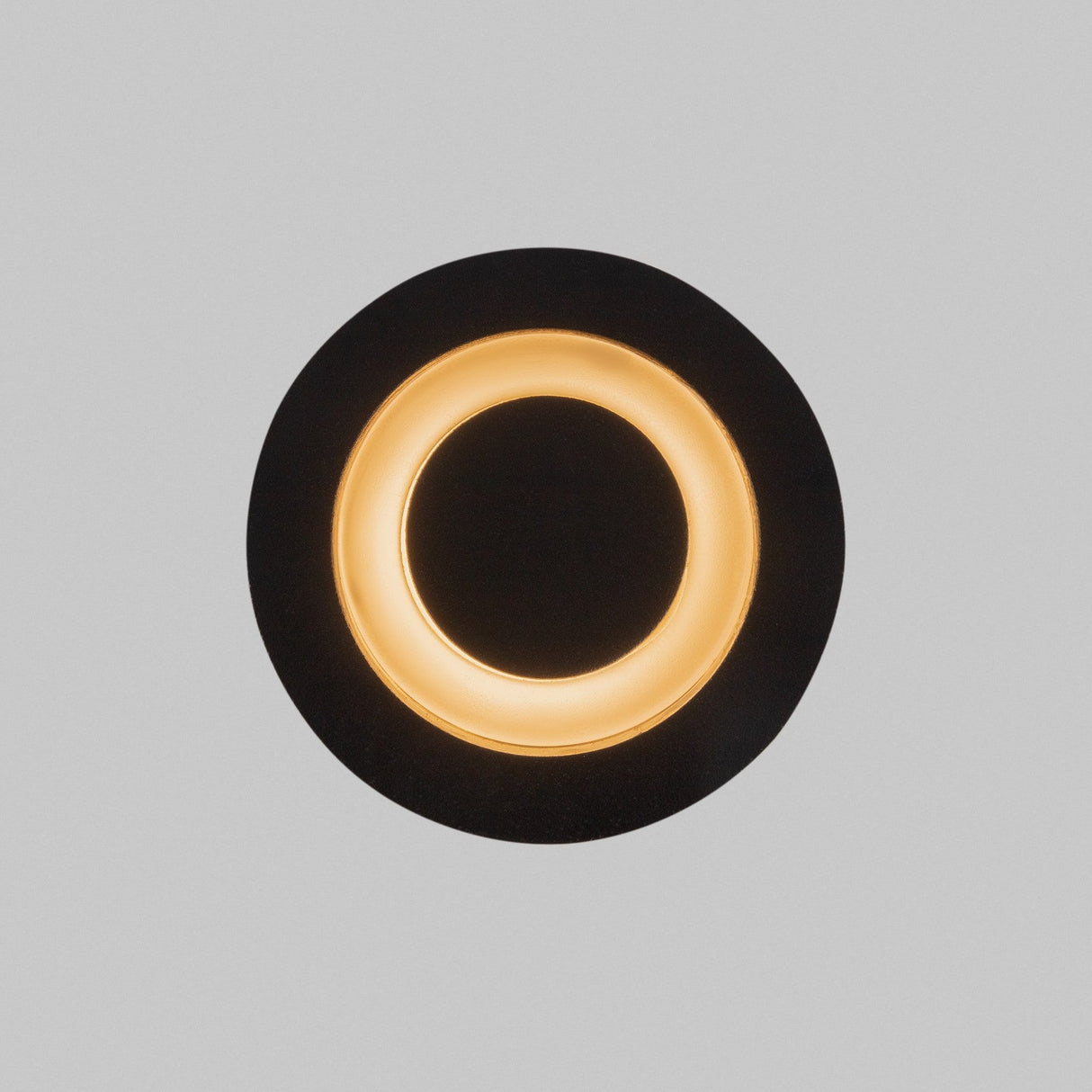 The Limo Outdoor LED Recessed Wall Light in black features a circular design with a glowing inner ring, emitting warm golden light. Its energy-efficient construction contrasts elegantly against a plain gray background, combining style and sustainability.