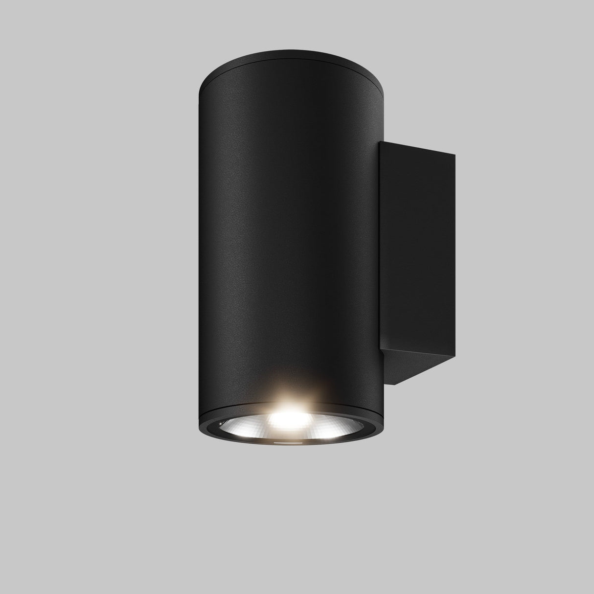 The Shim Outdoor Wall Light 10W 3000K in graphite is a modern cylindrical fixture elegantly mounted on a flat surface. Rated IP65, it illuminates downward, showcasing its minimalist design against a plain gray background.