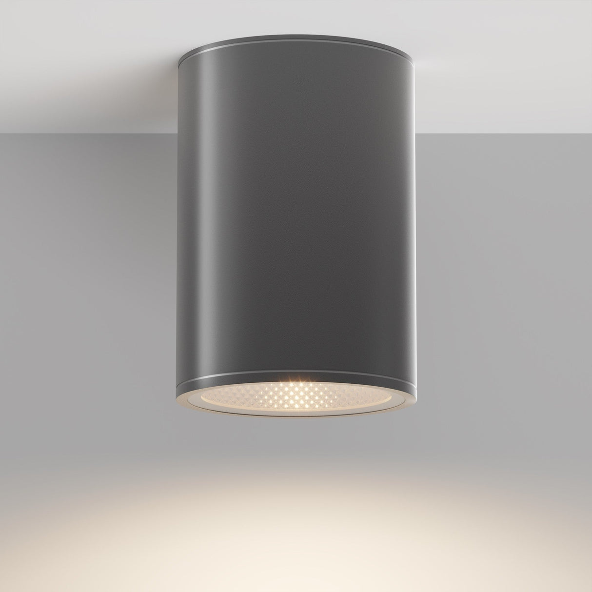 The Bar Outdoor LED Downlight 12W 3000K - Graphite, a minimalist cylindrical ceiling light with a graphite finish, is installed on a light gray ceiling. It emits an energy-efficient, soft warm glow downward to illuminate the area beneath.