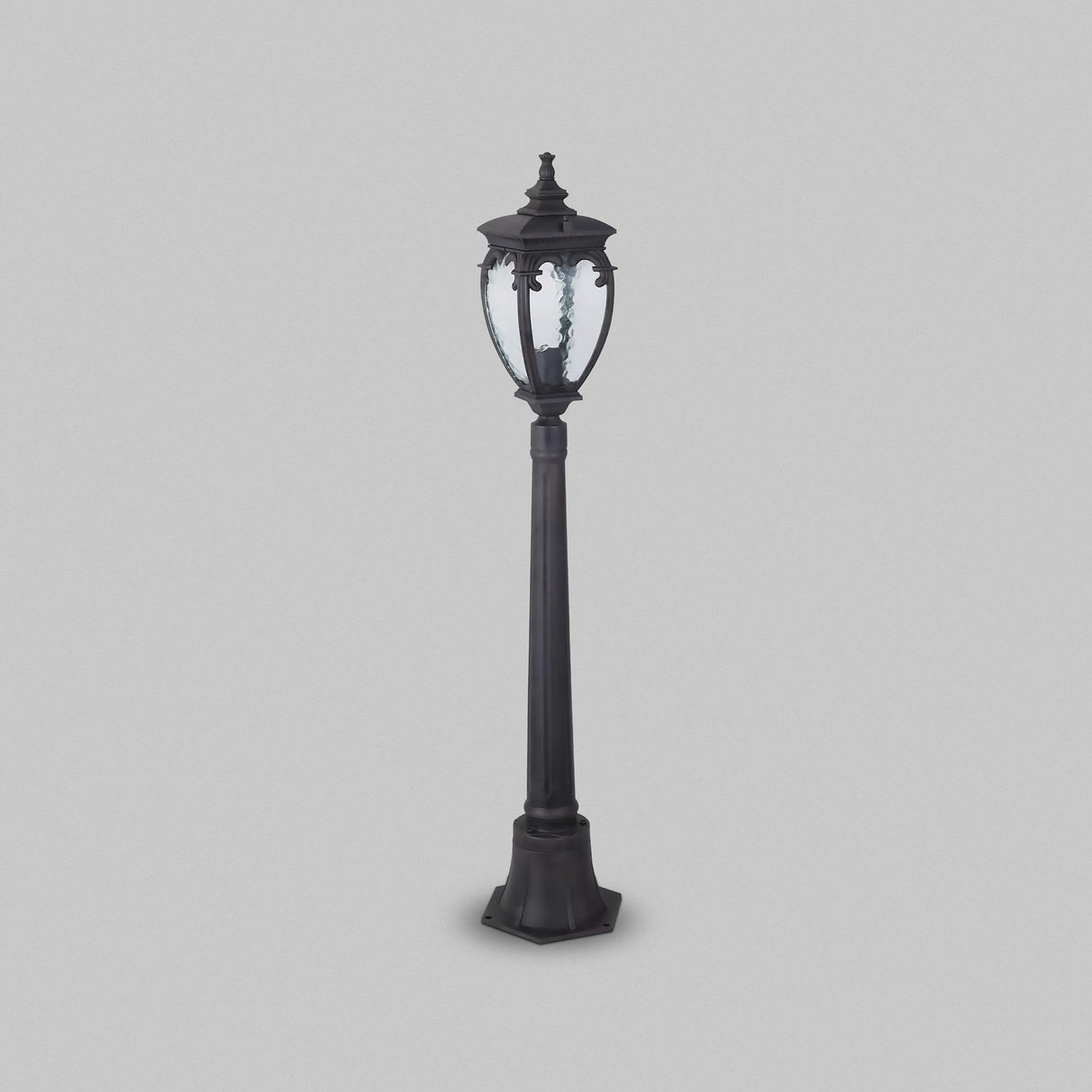 The Fleur 1 Light Pillar Lantern in bronze, designed with a vintage style and ornate detailing, features a clear glass lantern top. Made from weather-resistant aluminum, it’s perfect for outdoor lighting, set against a plain gray background.