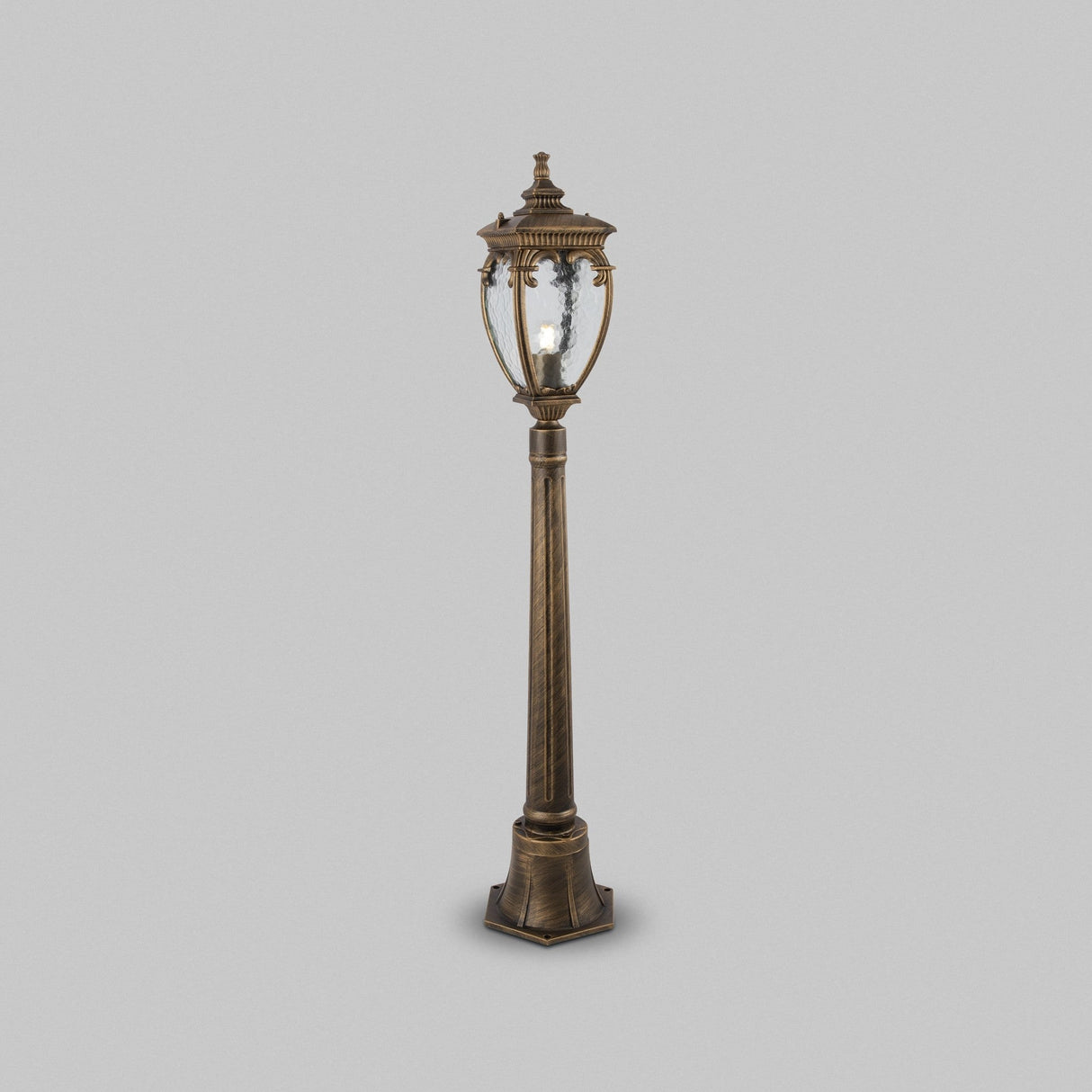 A vintage-style street lamp, Fleur 1 Light Pillar Lantern in black, with intricate detailing and a clear PULEGOSO glass lantern, stands against a plain gray background.