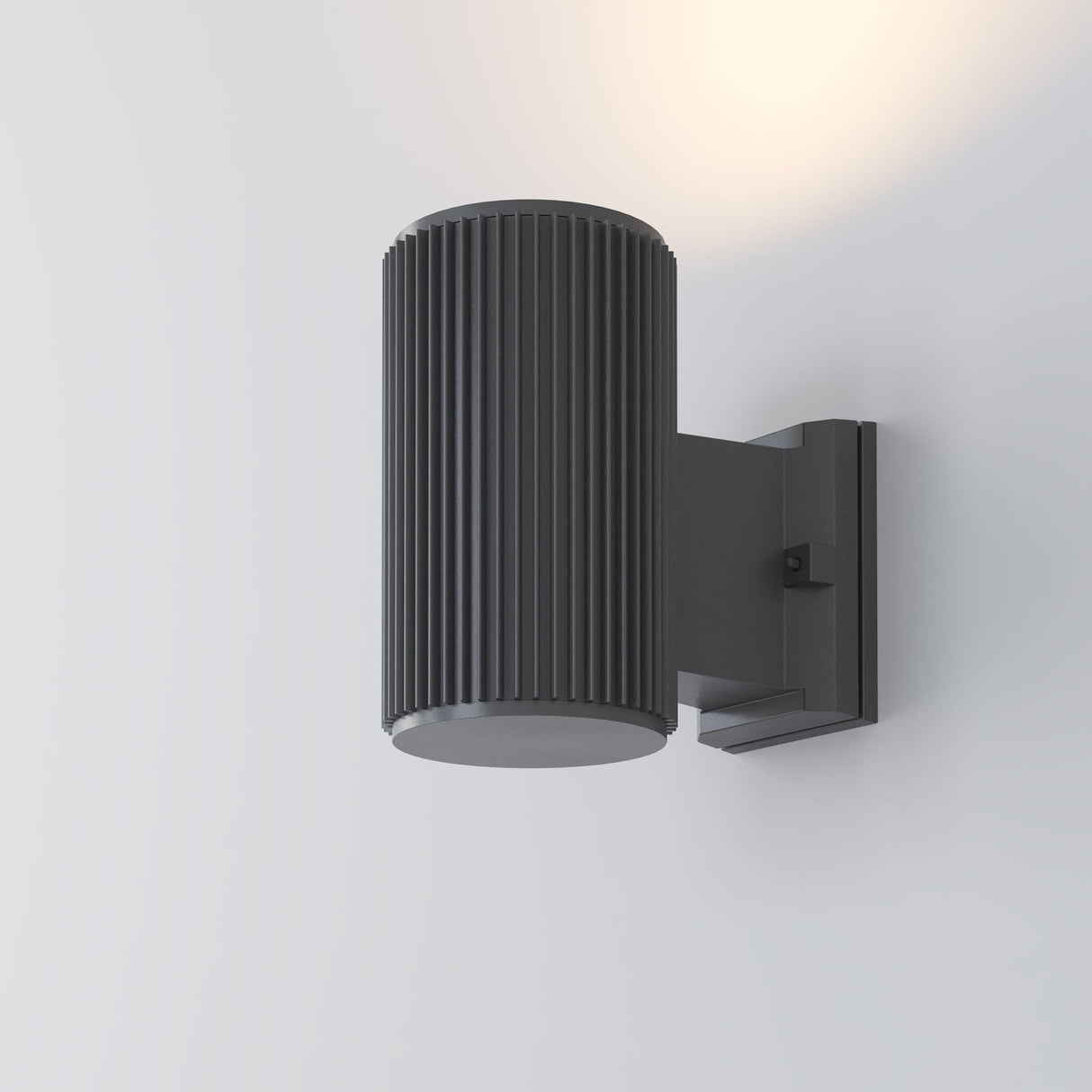 The Rando Outdoor Wall Light - Grey is a modern, cylindrical fixture with a ribbed texture. Mounted on a plain white wall, it features an IP54 rating and emits soft light from the top, enhancing its contemporary design with a subtle glow.