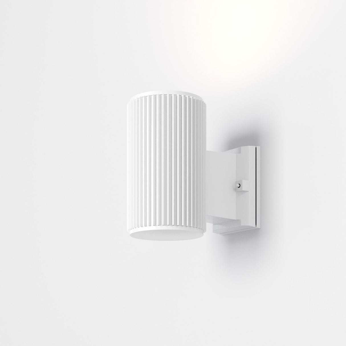 The Rando Outdoor Wall Light - White is a minimalist, wall-mounted cylindrical lamp with vertical grooves, emitting soft light. Designed for architectural lighting, it boasts an IP54 rating and attaches to a white rectangular base on the wall.