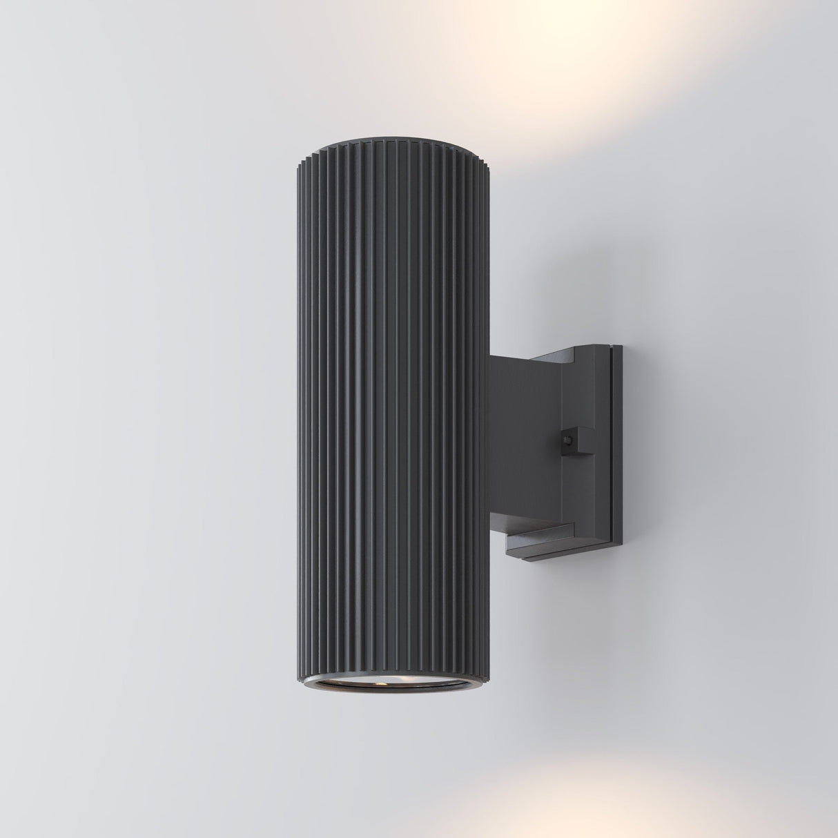 The Rando Outdoor Up & Down Wall Light - Grey features a ribbed grey design, casting dual-beam illumination upwards and downwards on a pale wall. It is corrosion-resistant for lasting durability.