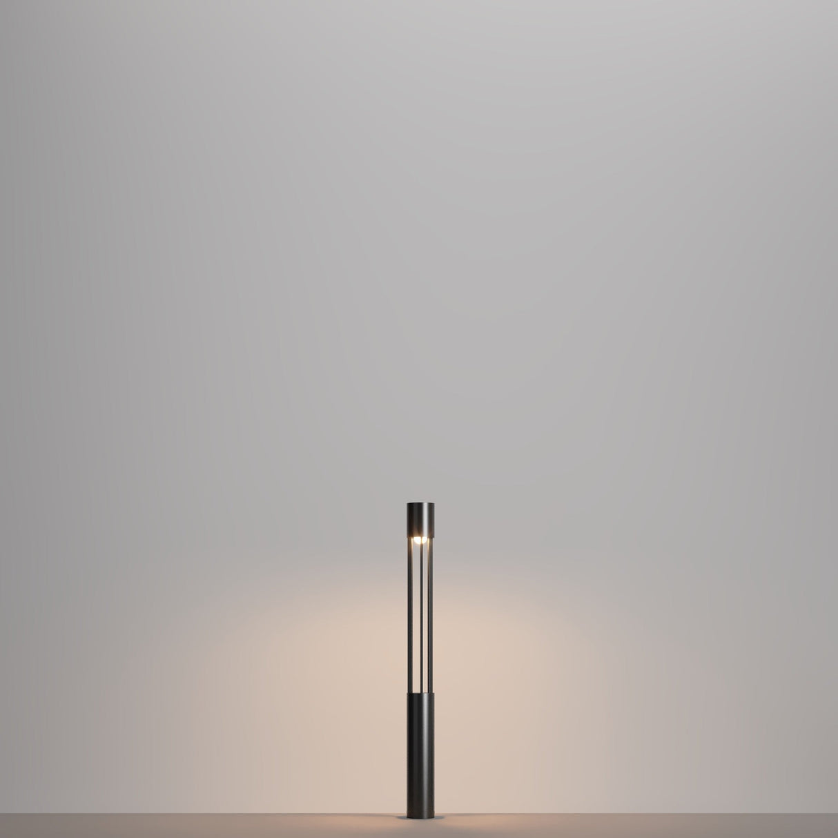 The Mill LED Short Bollard Light - Graphite, with its minimalist cylindrical black base and narrow vertical light tube, elegantly stands on a plain surface. Its graphite finish enhances the soft glow on the neutral gray wall behind it.