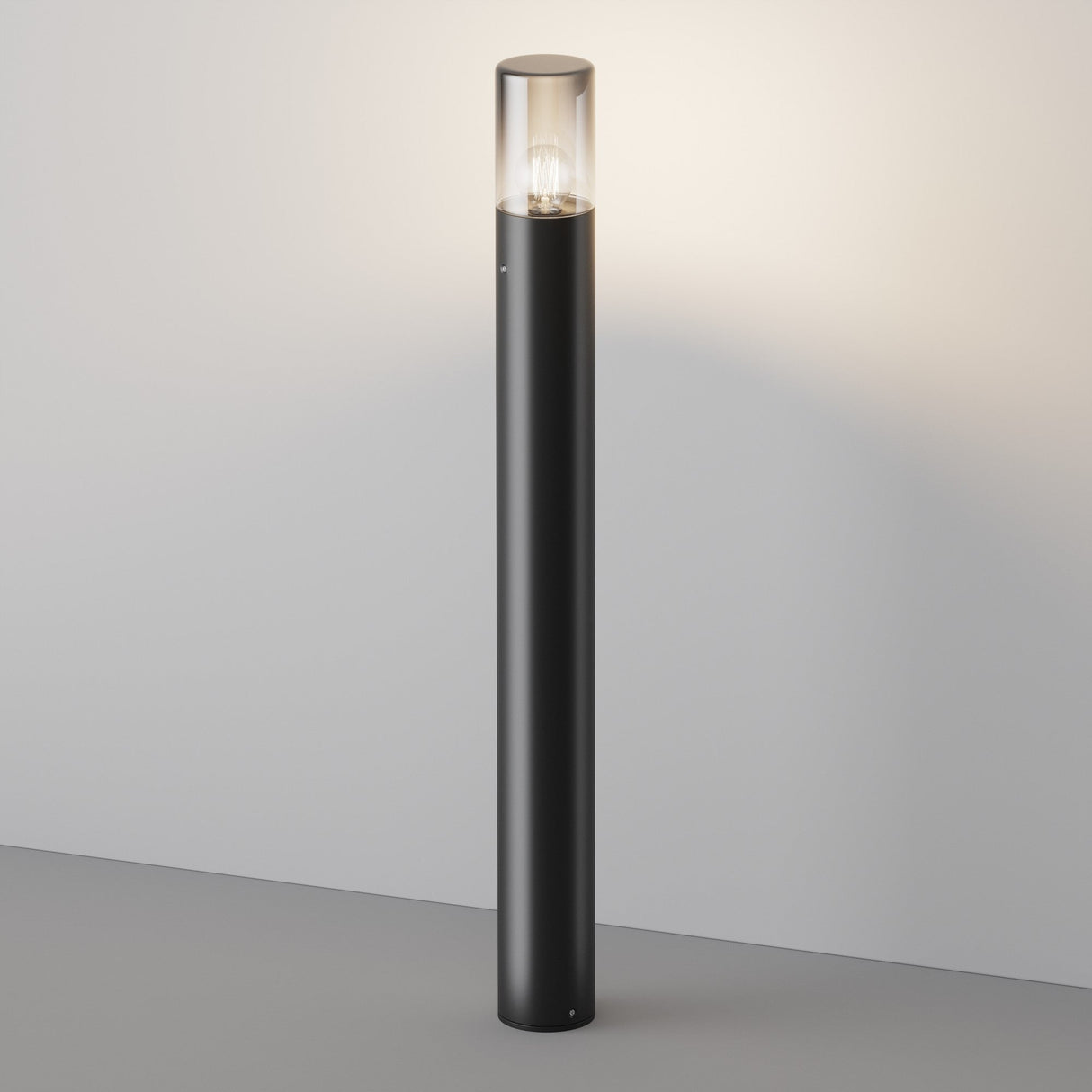 The Amas Bollard Light 80cm in sleek graphite features a modern cylindrical design and transparent top revealing a glowing bulb. It stands elegantly on a gray surface, casting a soft glow onto the wall, perfect for outdoor ambiance.