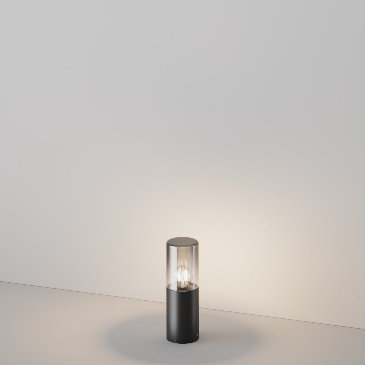 The Amas Bollard Light 25cm in Graphite emits a soft, warm glow that illuminates a plain white surface and casts a gentle glow on the wall, offering ambient light with its sleek, cylindrical design.