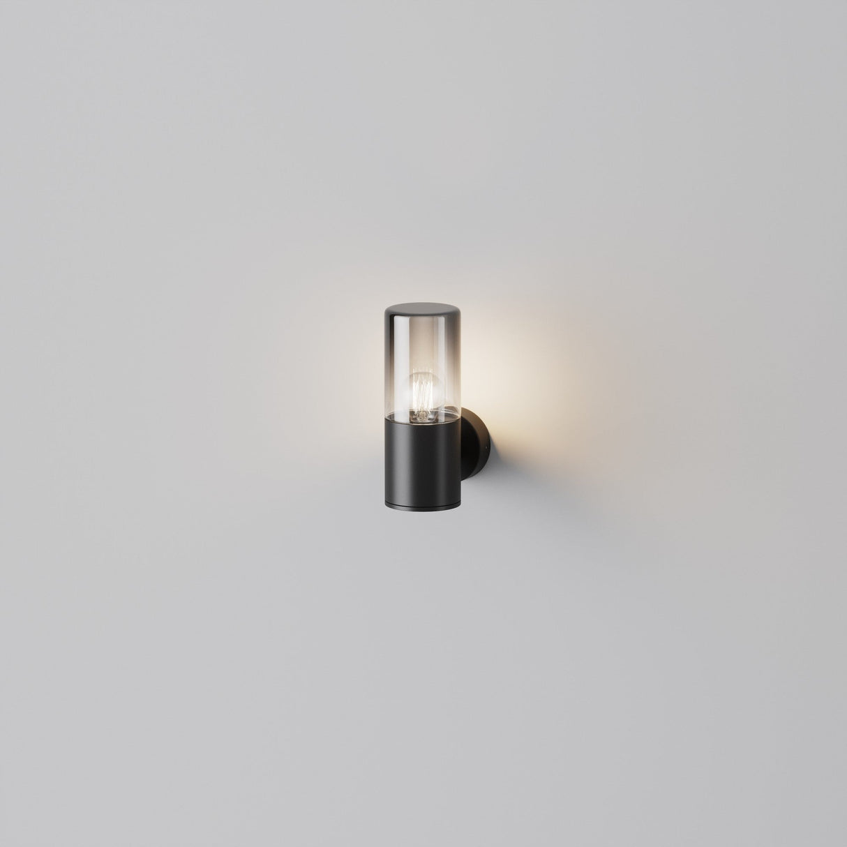 The Amas Outdoor Wall Light - Graphite features a modern, weather-resistant aluminum cylinder with a chic graphite lower half and a clear upper section that reveals the bulb, elegantly casting a soft glow on surrounding white walls.