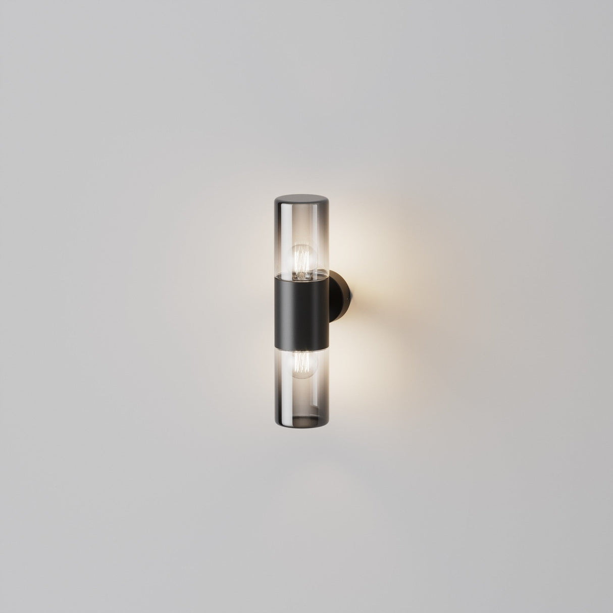 The Amas Outdoor Up & Down Wall Light - Graphite features a modern cylindrical glass design with two visible bulbs on a graphite finish base. Its weather-resistant construction emits a warm glow against a plain white wall.