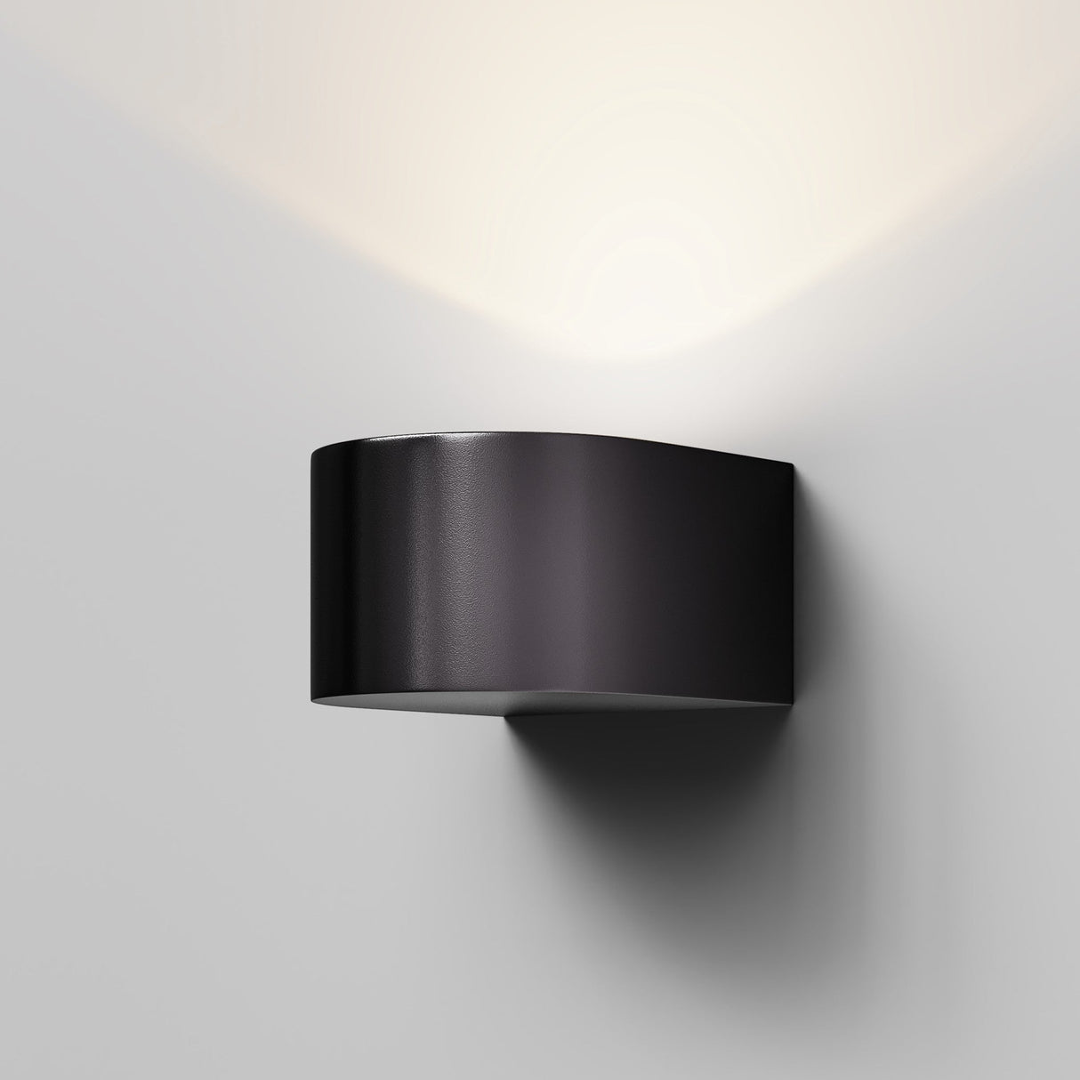 The Blow Outdoor LED Wall Light in Graphite, a modern and minimalist aluminum fixture, is mounted on a white wall. Its energy-efficient design projects an upward triangular glow above the fixture.