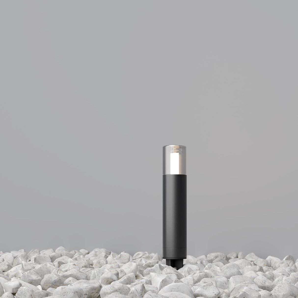The Glide Outdoor Short Spike Light in Graphite is a modern cylindrical black lamp with a clear top. It emits light while standing on white stones against a gray background, serving as an elegant pathway marker lighting solution.
