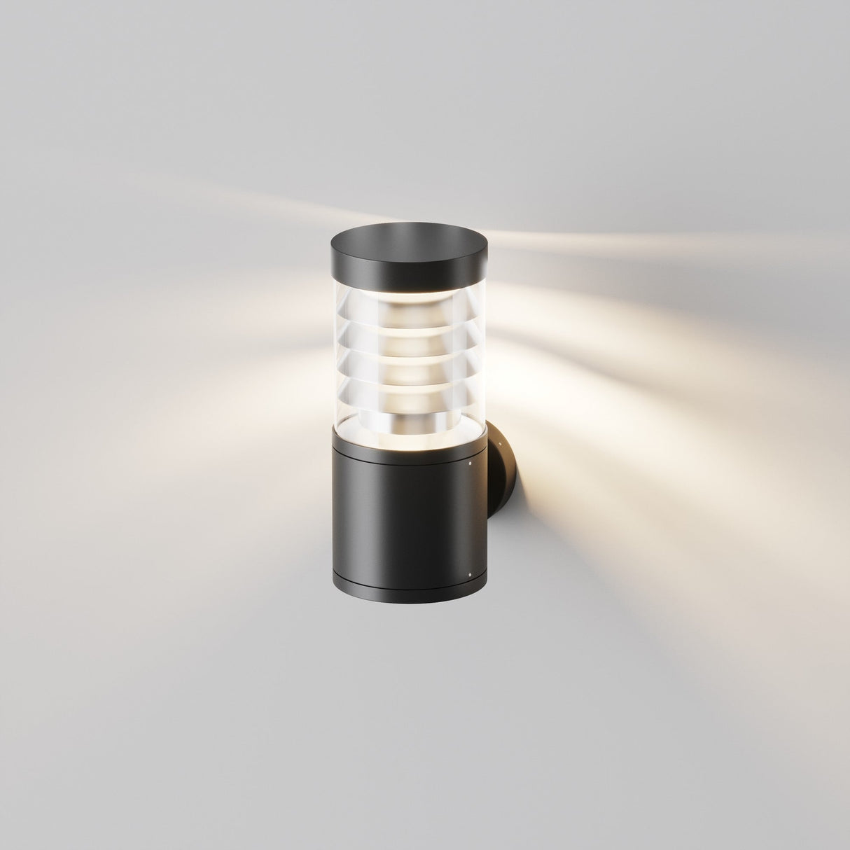 The Spir Outdoor Wall Light 3000K 12W in graphite is a modern black sconce with a cylindrical design, featuring a layered translucent section. It emits a soft glow that creates an enchanting pattern on the wall.