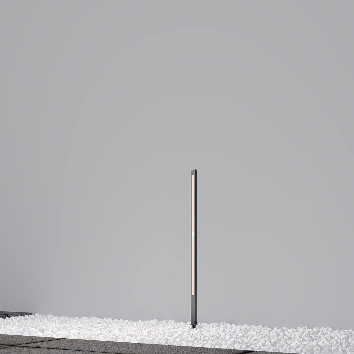 A minimalist outdoor scene includes the Pole LED Outdoor Spike Light 80cm in Graphite, a slim vertical fixture with an IP65 rating, standing on white pebbles against a pale gray wall.