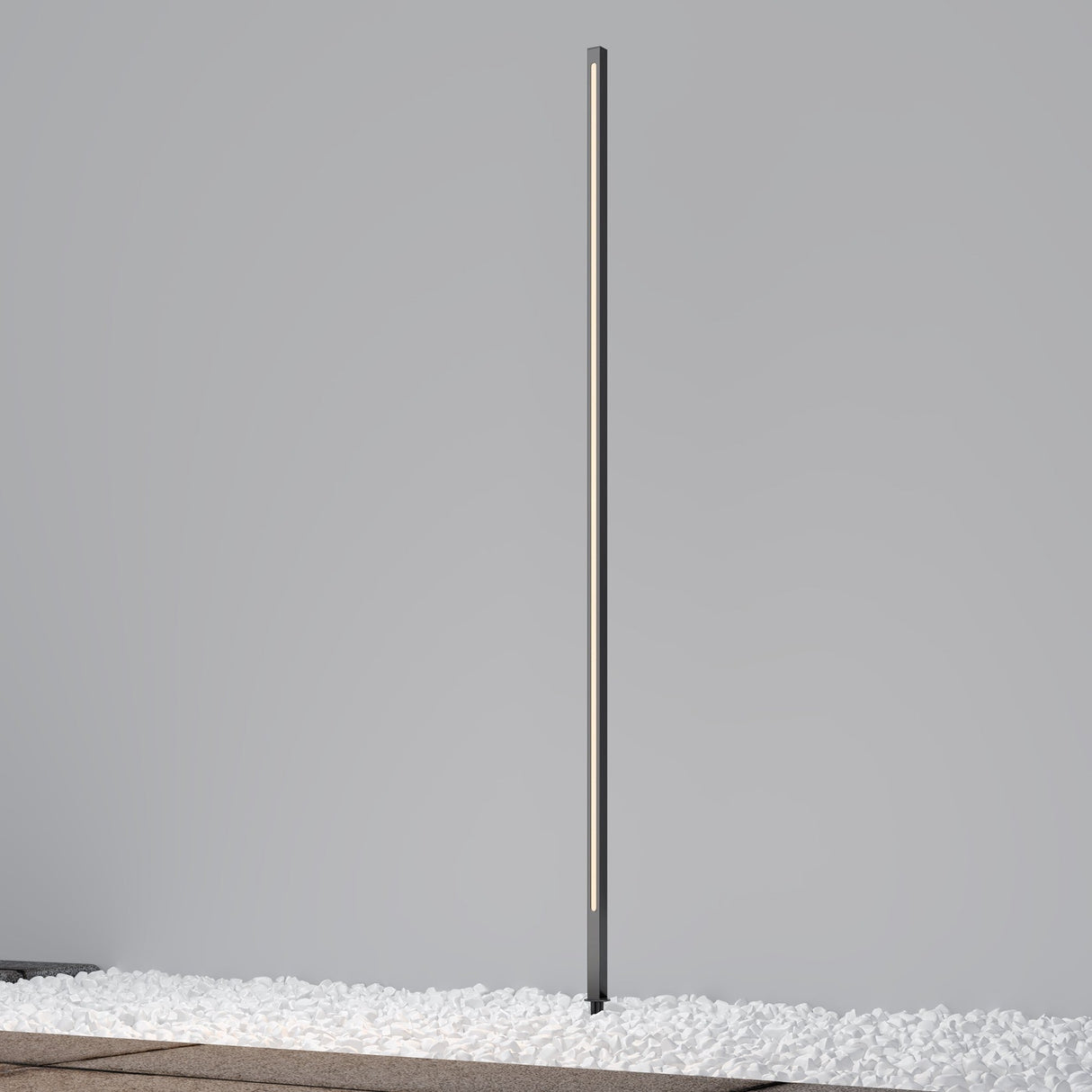 A tall, slim Pole LED Outdoor Spike Light 160cm - Graphite stands vertically against a plain gray background. Set on a floor covered with white pebbles, its turned on, emitting a soft, even glow ideal for outdoor lighting.