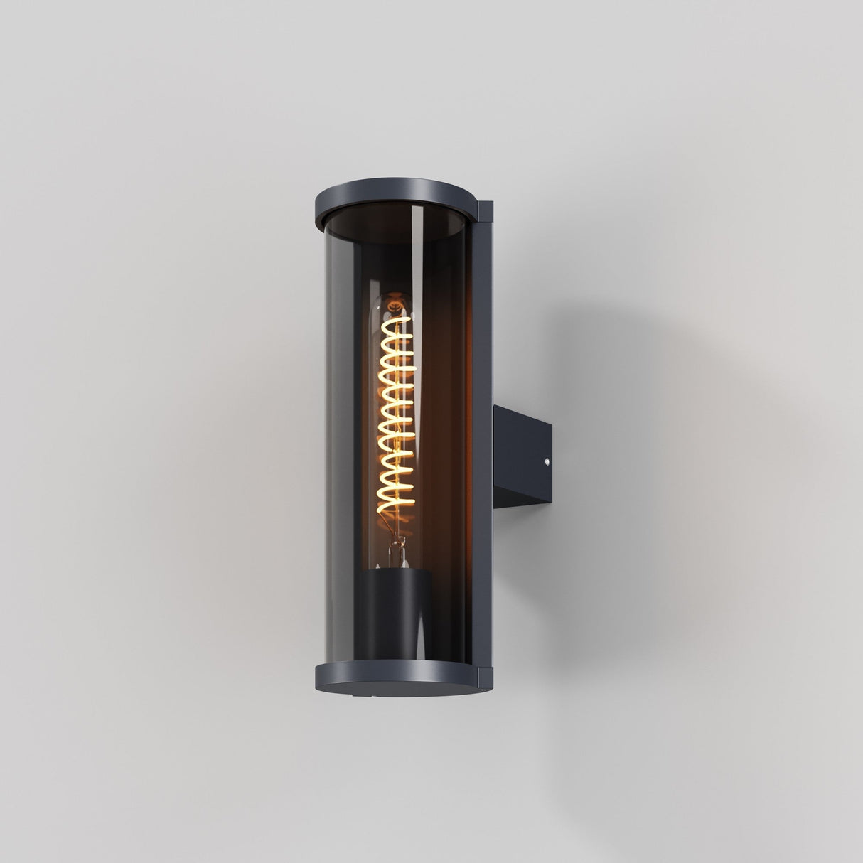 The Conf Outdoor Round Wall Light - Graphite offers a modern cylindrical design with IP54 protection. It features a glowing spiral filament inside transparent glass, mounted on a wall with a black metal bracket. Ideal for contemporary exteriors against smooth, light gray backgrounds.