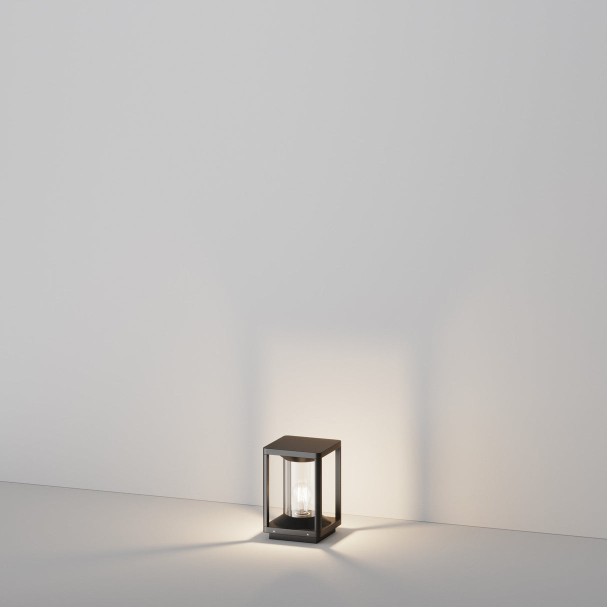 A 22cm Cell Pedestal Light in graphite, with a glowing bulb inside, sits on a plain white surface. Its soft illumination highlights outdoor elegance against the minimalist white wall backdrop, echoing an IP54 rating for durability.