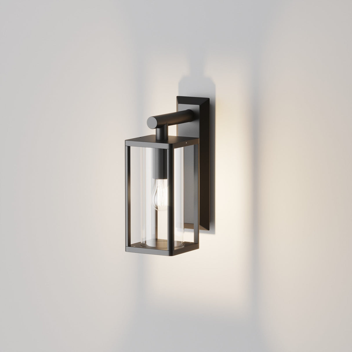 The Cell Outdoor Down Wall Light - Graphite features a sleek design with a black metal frame and clear glass panels. Its IP65 rating guarantees durability in diverse weather conditions, while the visible bulb provides a gentle glow, casting delicate shadows on the wall.