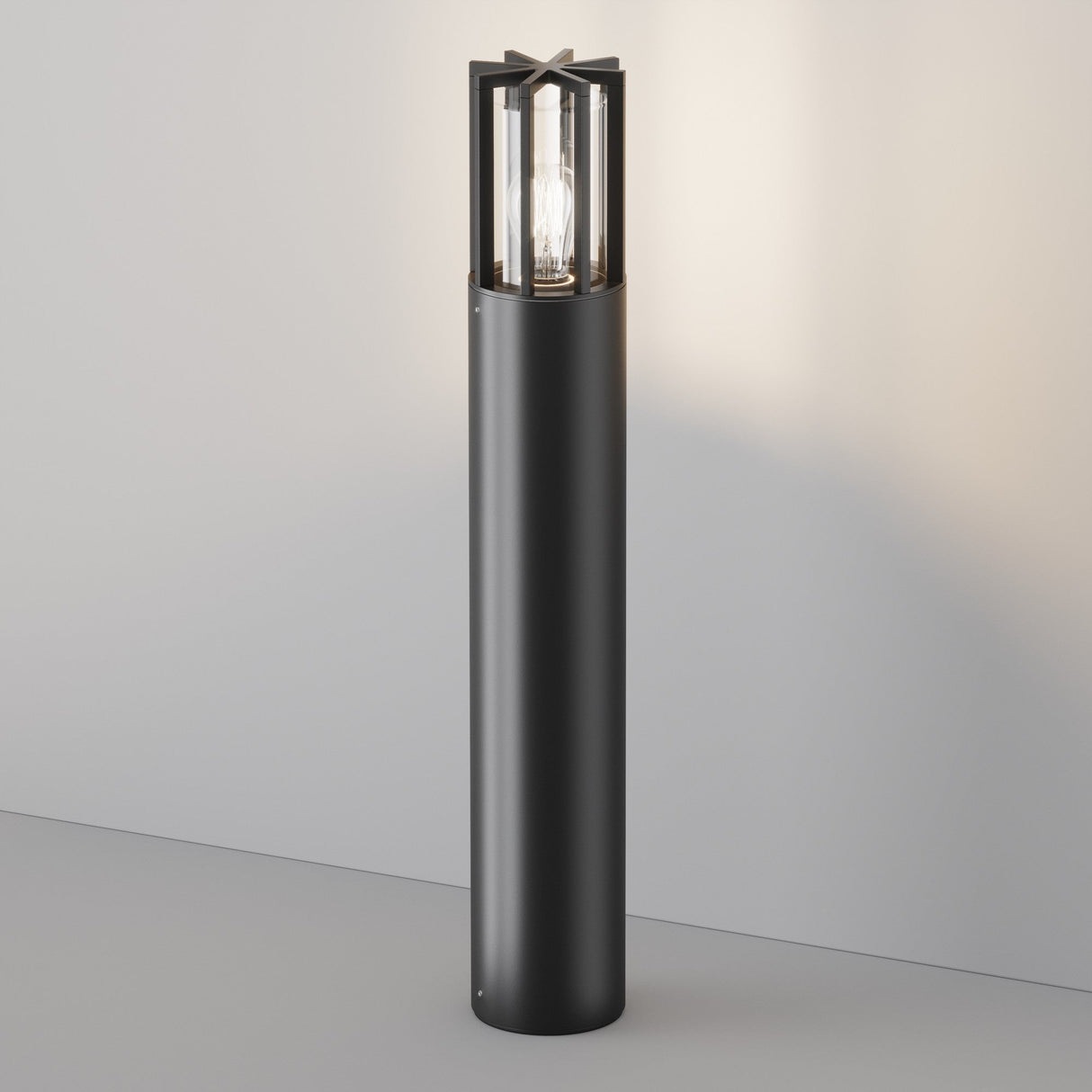 The Barrel Bollard Light 75cm in Graphite, a contemporary black cylindrical lamp with a visible bulb encased in glass and metal, casts a soft glow. Positioned against a light wall, its perfect for indoor elegance and outdoor lighting sophistication.