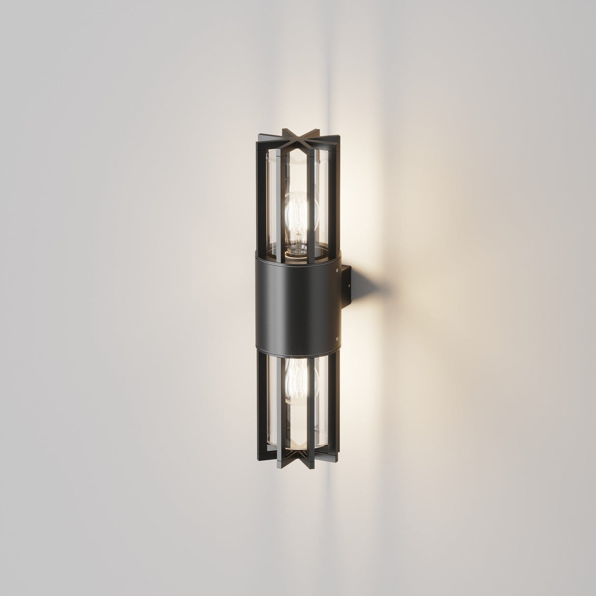 The Barrel Outdoor Up & Down Wall Light - Graphite, featuring a black metal frame with glass panels and two exposed bulbs, is mounted on a light gray wall. It casts a soft glow with sharp angles and symmetry, perfect for contemporary outdoor spaces.
