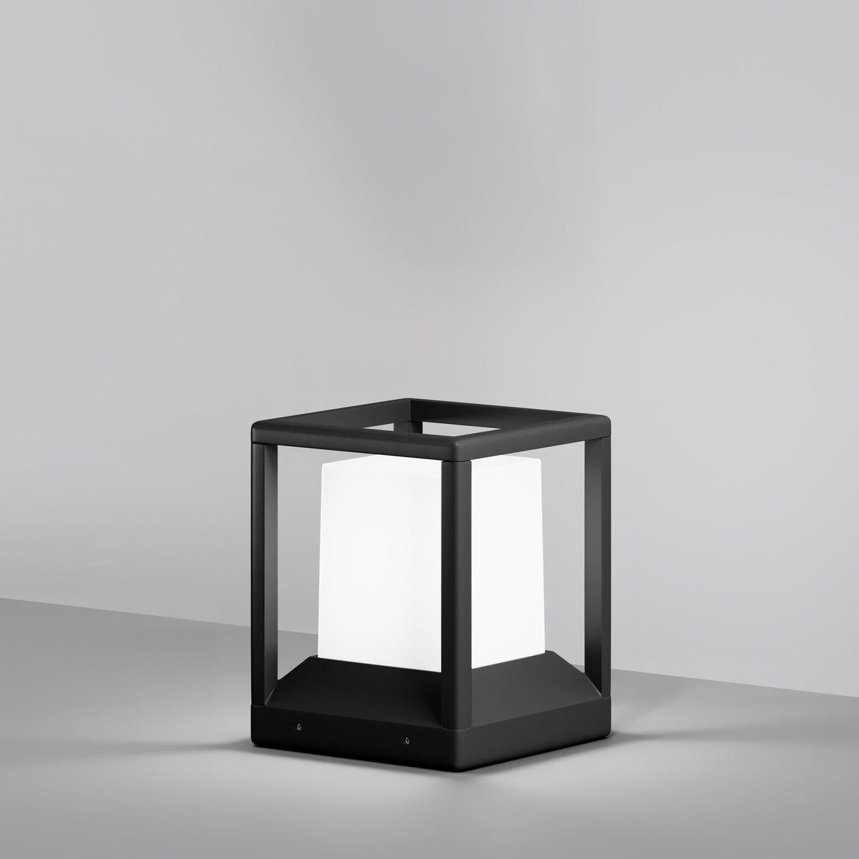 The Trim Pedestal Light 210mm in Graphite is a modern, minimalist table lamp with an aluminum body. It features a black square frame and a glowing white cube light at the center, set against a plain gray background.
.