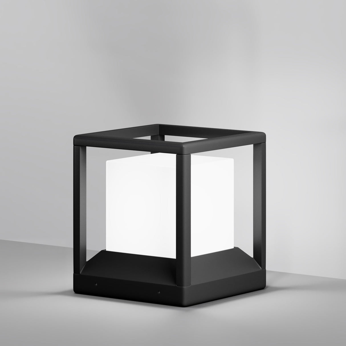 The Trim Pedestal Light 255mm in graphite boasts a modern black cubic design with a glowing white cube centerpiece on a neutral gray surface. Its minimalist open frame and sleek, contemporary look make it perfect for outdoor lighting against a simple, light-colored wall.