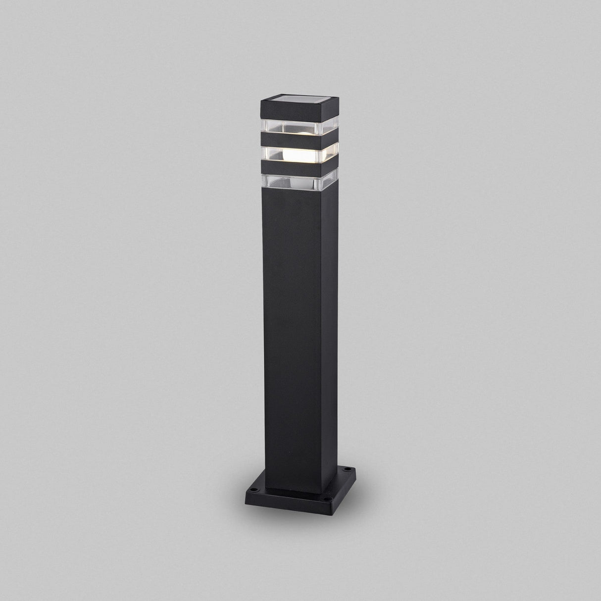 The Unter den Linden Bollard Light - Black is a modern, rectangular outdoor light featuring a sleek black base and a transparent top section for illumination. Built with corrosion-resistant materials for durability, it showcases horizontal lines against a plain gray background.