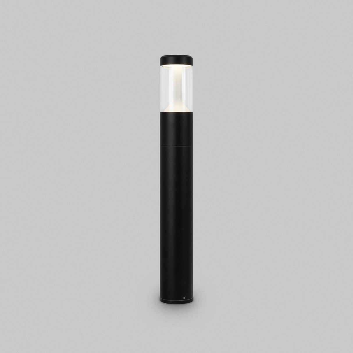 The Koln LED Bollard Light 65cm - Black is a tall, black cylindrical outdoor fixture with a transparent top section and an efficient LED module, casting a warm glow against a plain gray background.