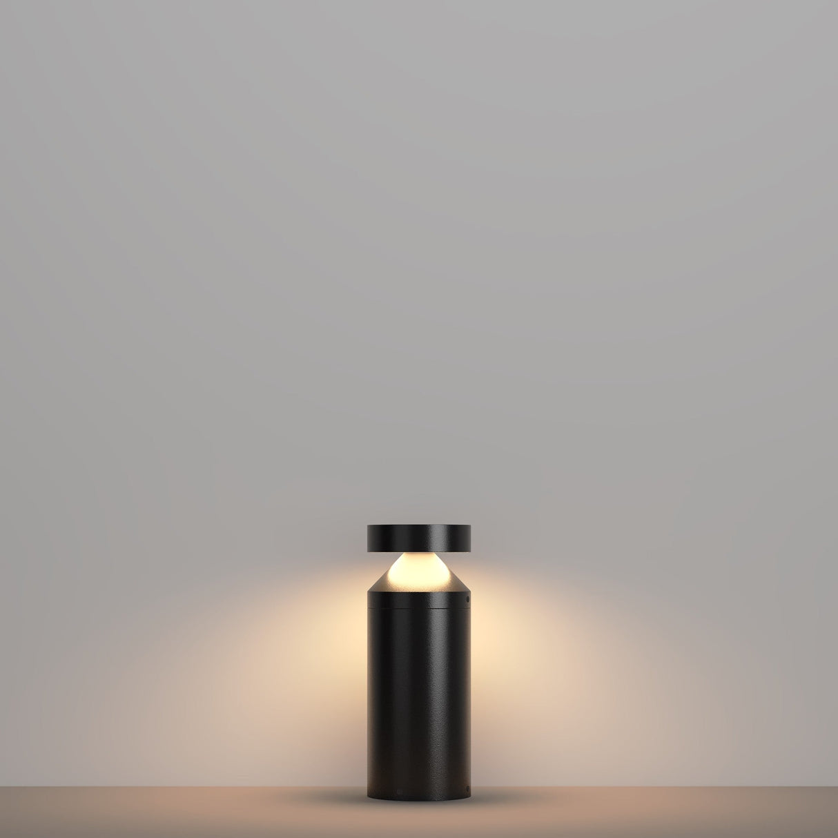 A minimalist black table lamp with a cylindrical base and flat top rests on a grey surface. Its energy-efficient design mirrors the Olten Bollard Light 30cm in Graphite, emitting a warm glow against the grey background for a calm, cozy ambiance.