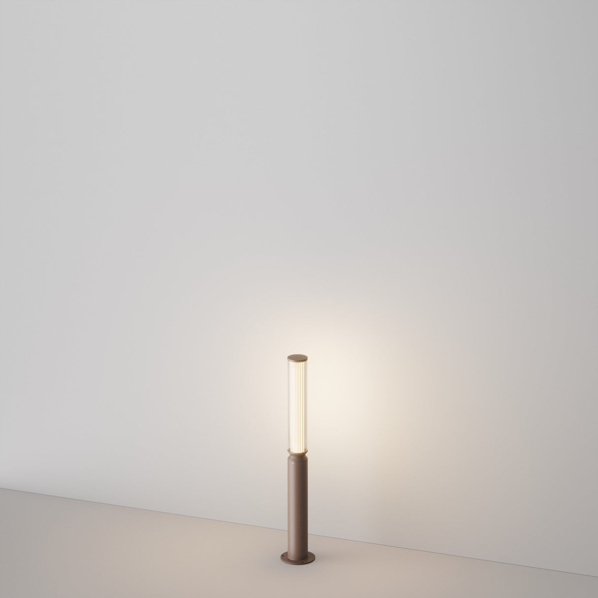 The Lit LED Short Bollard Light - Brown, a minimalist cylindrical lamp, emits a warm, energy-efficient glow on a smooth light-colored floor and casts gentle illumination against a plain, light wall.