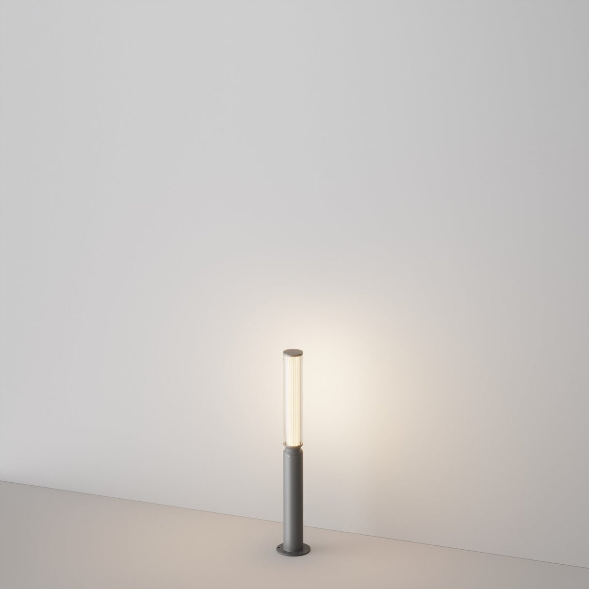 The Lit LED Short Bollard Light in a modern graphite finish casts soft light against a light-colored wall, with its minimalist design and muted gray base on a smooth surface creating a calm, outdoor-like ambiance.