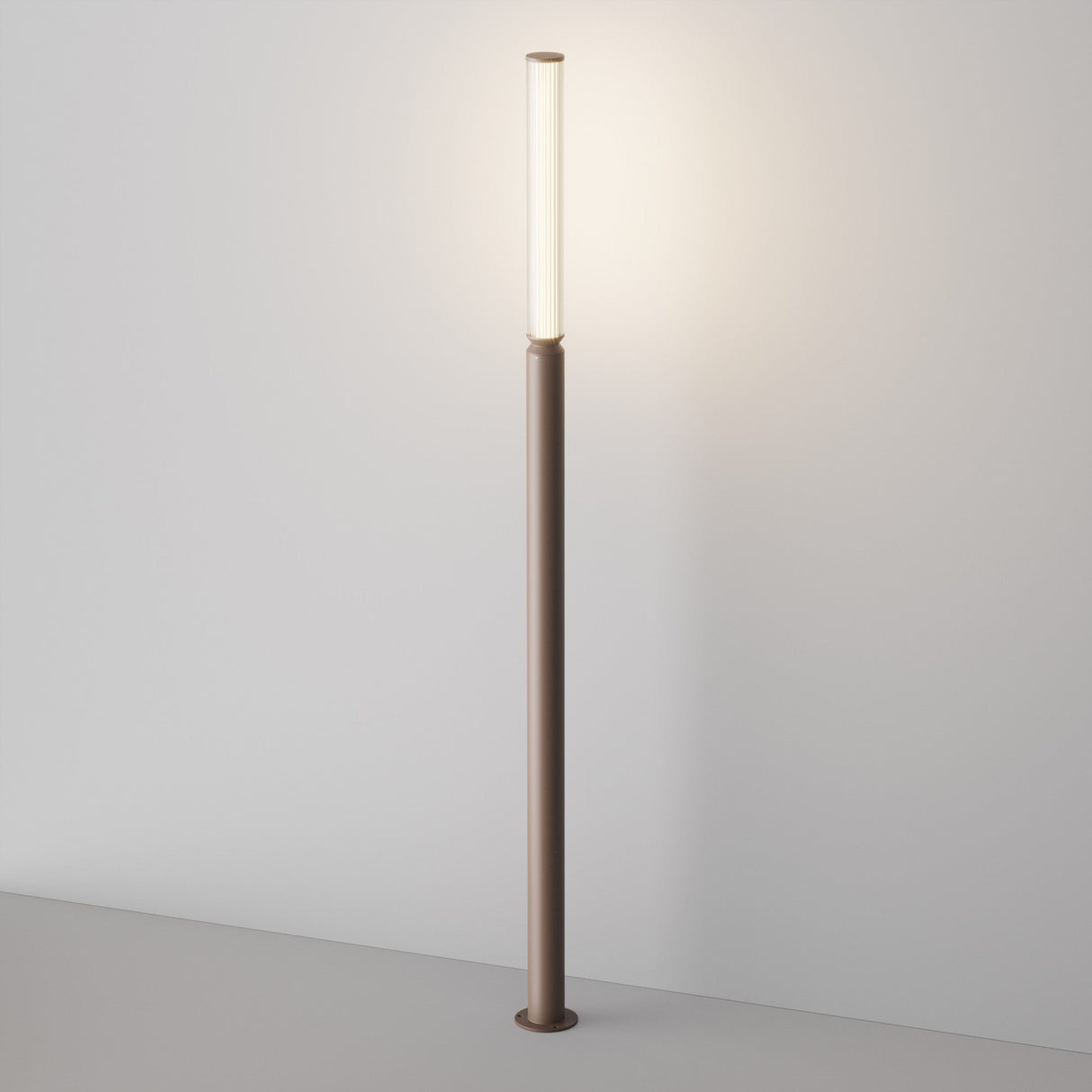 The Lit LED Tall Bollard Light - Brown is a modern, slim floor lamp with energy-efficient lighting and a cylindrical top. Its minimalist design casts a soft glow against white walls, featuring a sleek brown base and pole for a contemporary look.