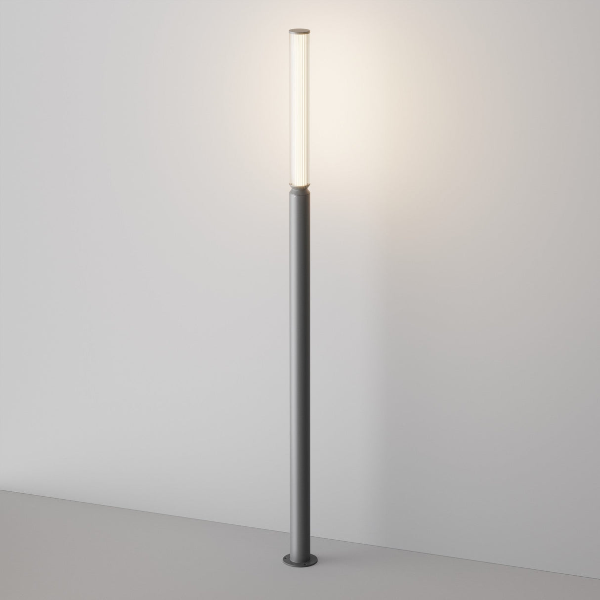 The Lit LED Tall Bollard Light - Graphite is a modern floor lamp with an energy-efficient system, featuring a tall cylindrical pole and slim vertical light source emitting a warm glow, standing against a plain white wall on a light gray floor.